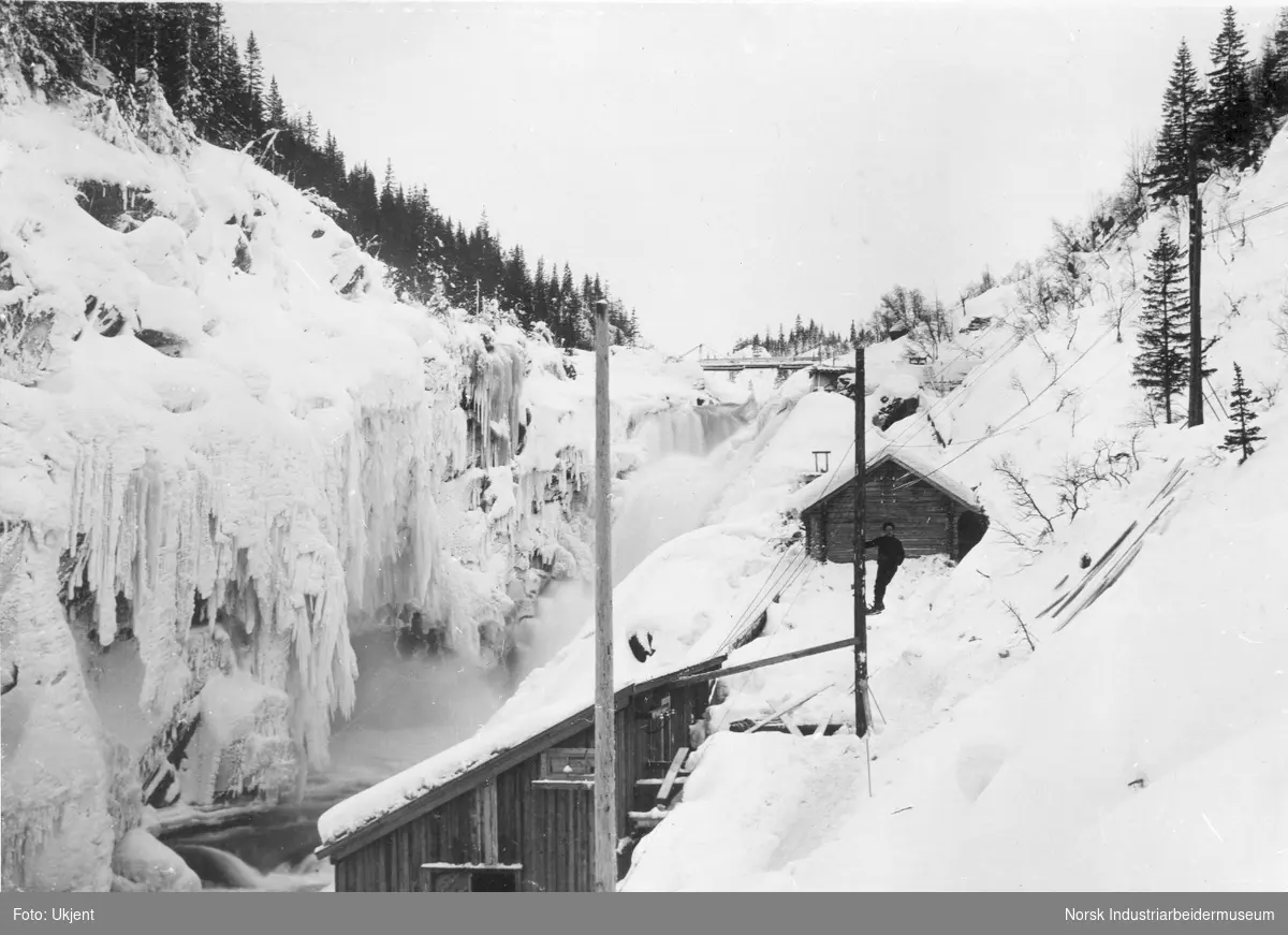 Mann stående i strømmast ved 300 H.K. kraftstasjon i juvet mellom Krokan og Rjukanfossen bygget i 1907. På oversiden "Dynamohuset" som ble bygget i 1897. Broen over til fordelingsstasjonen på Vemork mot vest.
