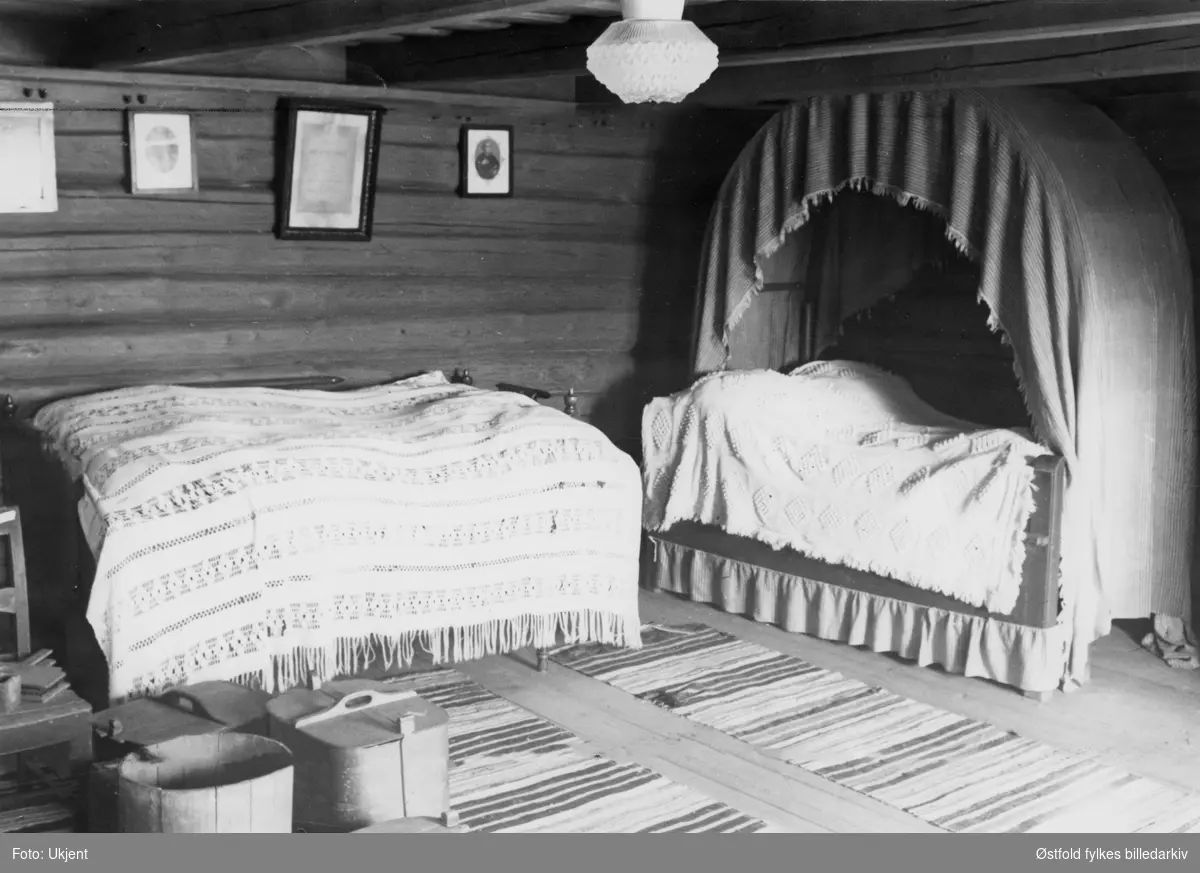 Gården Alvim nordre i Tune, fotografert februar 1977.
Fra våningshus, soveromsinteriør - ant fra kammers eller loftsrom. Seng (uttrekksseng?) med teppe,omhengsseng, tiner,  bilder, lampe, filleryer - vevde.
Våningshus bygd omrking 1780, restaurert 1955.