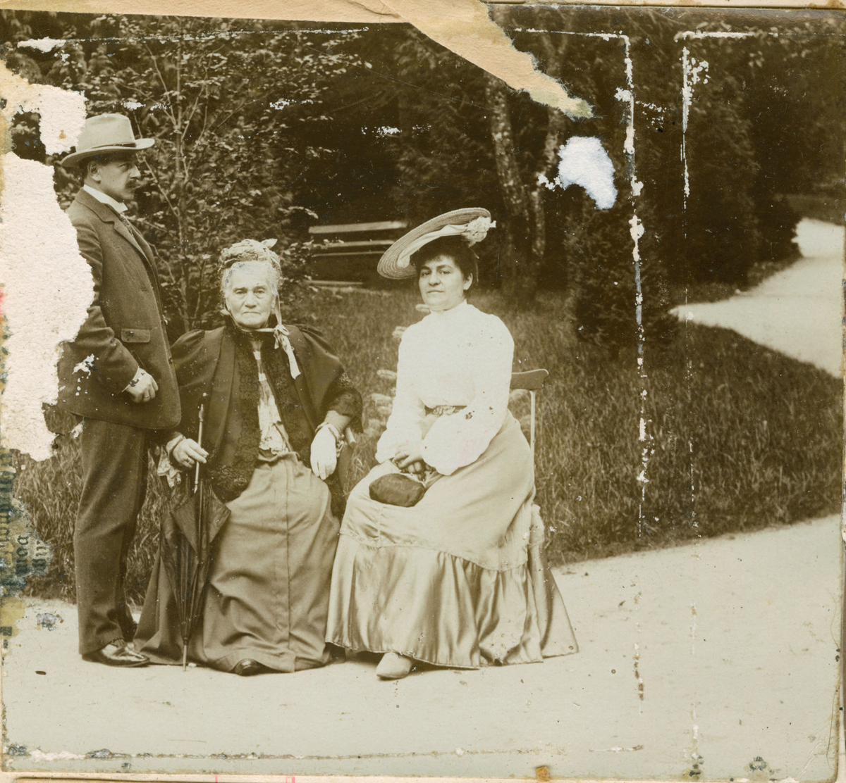 Motivet viser en mann og to kvinner fotografert utendørs i en park. Mannen står vendt mot de to sittende kvinnene. Den eldste kvinnen holder en paraply og bærer et hodeplagg knyttet under haka. Den andre kvinnen hviler en poseveske på fanget.