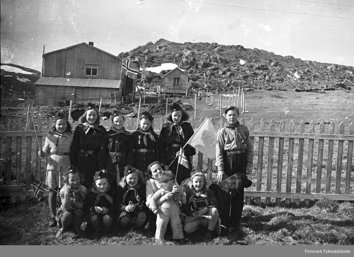 Gruppebilde av speidertropp Havøysund 1950. Eli Bjørnstad foran. En hund. Bildet tatt antakelig i 17. mai. Norsk flagg i bakgrunn.