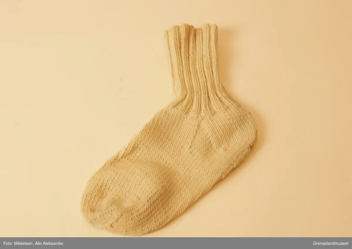 Gjenstanden er en enslig hvit sokk av håndstrikket bomull. Vrangbord to rette, to vrange. Stoffet er litt stivt, men ser relativt ubrukt ut.