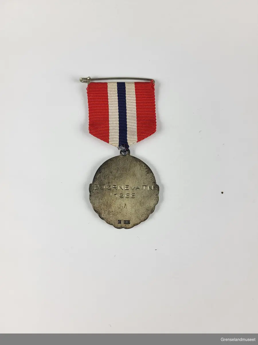 En lyre harpe i sølv uthevet fra resten av sølvmedaljen. Det norsk flaggets farger i tekstil som er festet til medaljen.  