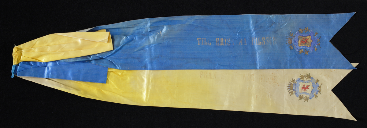 Blått och gult hyllningsband i siden med förgylld tryckt skrift; "Till Christina Nilsson. Från Smålands gille i Göteborg 18 15/9 85".
På bandändarna Smålands vapen.

Inskrivet i huvudkatalog 1933.