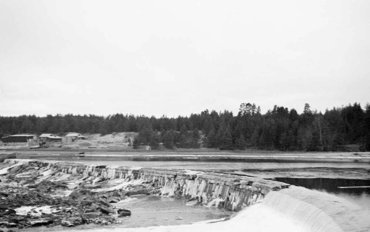 Fra dammen som stuvet opp vann til kraftverket ved Skjefstadfossen i Glomma, i Heradsbygda i Elverum. Fotografiet ble tatt høsten 1939, og det er montert som den høyre delen av et panoramabilde i et av Glomma fellesfløtingsforenings album (jfr. SJF.1989-03535). Her ser vi overløpsdammen, som var 312 meter lang, og litt lavere i den østre enden (til høyre på dette bildet) enn i den vestre. Denne løsningen ble valgt for å skape et drag i vannet som trakk fløtingsvirket mot den østre delen av elveløpet, der det var noe djupere enn i den midtre og vestre delen, og følgelig noe mindre fare for at stokkene skulle sette seg fast og pakkes sammen i hauger som det ble krevende og farlig for fløterne å løsne.