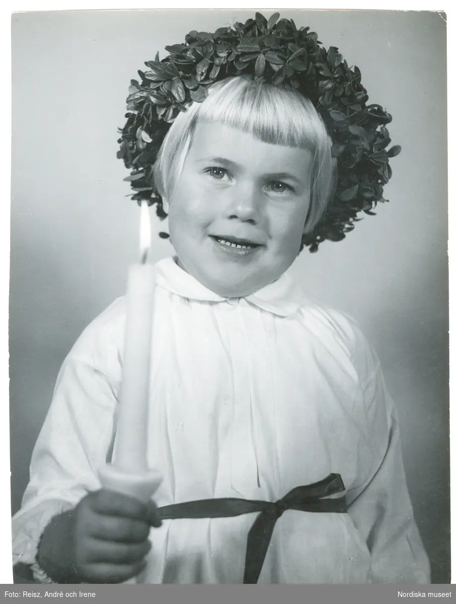 Porträtt av en flicka klädd i Luciaklänning och med krans på huvudet, band om midjan och ett ljus i handen.