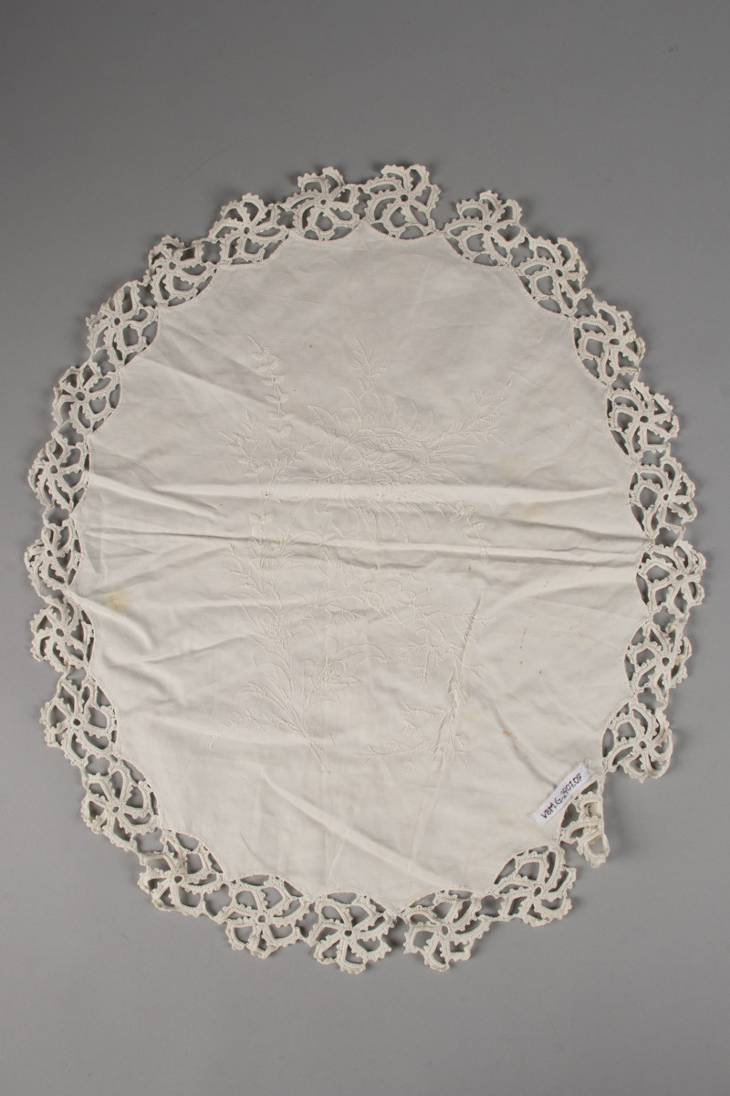 Oval, nesten rund duk med brodert blomstermotiv, hvit tråd på hvit tekstil. Heklet blondekant. Noen flekker.