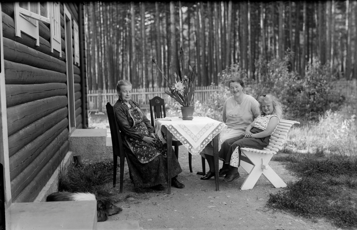 Gruppebilde.Ved et bord i hagen sitter mor til Alma,Alma og Gard Langslet. Fin duk på bordet.
