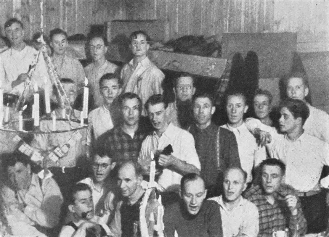 Fotografi av julefeiringa på Grini 1944. Flere mannlige fanger med ansiktet vendt mot kamera. I midten ser man Otto Nielsen med ukulelen i hånda.
