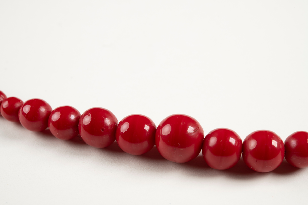 Kort halsband av röda plastpärlor trädda på en nylonlina. En stor pärla i mitten och sedan successivt mindre pärlor på ömse sidor fram till springring och ögla i metall som utgör halsbandets lås. Nylonlinan knuten i låset.