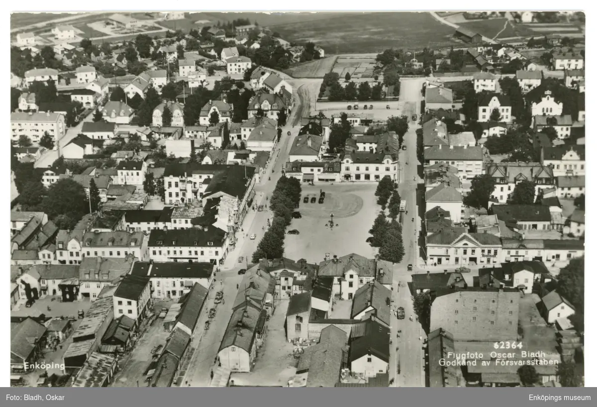 Vykort med flygfoto över de centrala delarna av Enköping. Det parallella gatorna i bild är från vänster: Källgatan och Torggatan. I mitten av bilden ser vi stora torget. Vi ser även att både busstationen, Gustaf Adolf plan, Sparbanken och "Funkisvillan" är klara vilket innebär att bilden fotograferades före 1943.