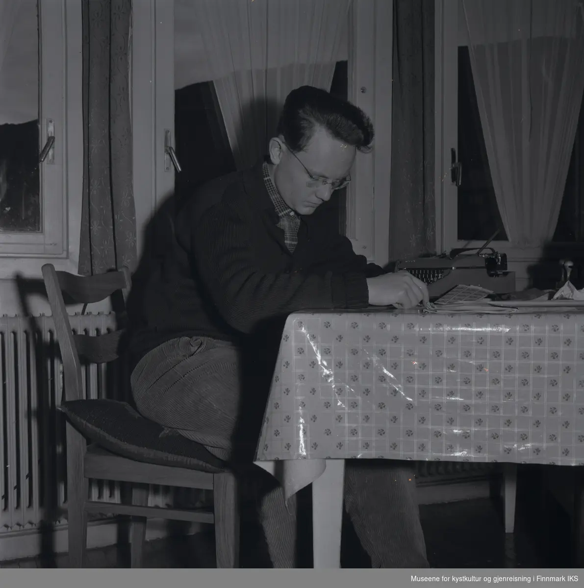 Friedrich Hörl sitter ved et bord og leser. På bordet, som er dekket med en voksduk, er det plassert en skrivemaskin, stempler og frimerker, samt en avis. Den unge mannen har en brille og er ikledd fløyelsbukse, rutet skjorte og strikket jakke.