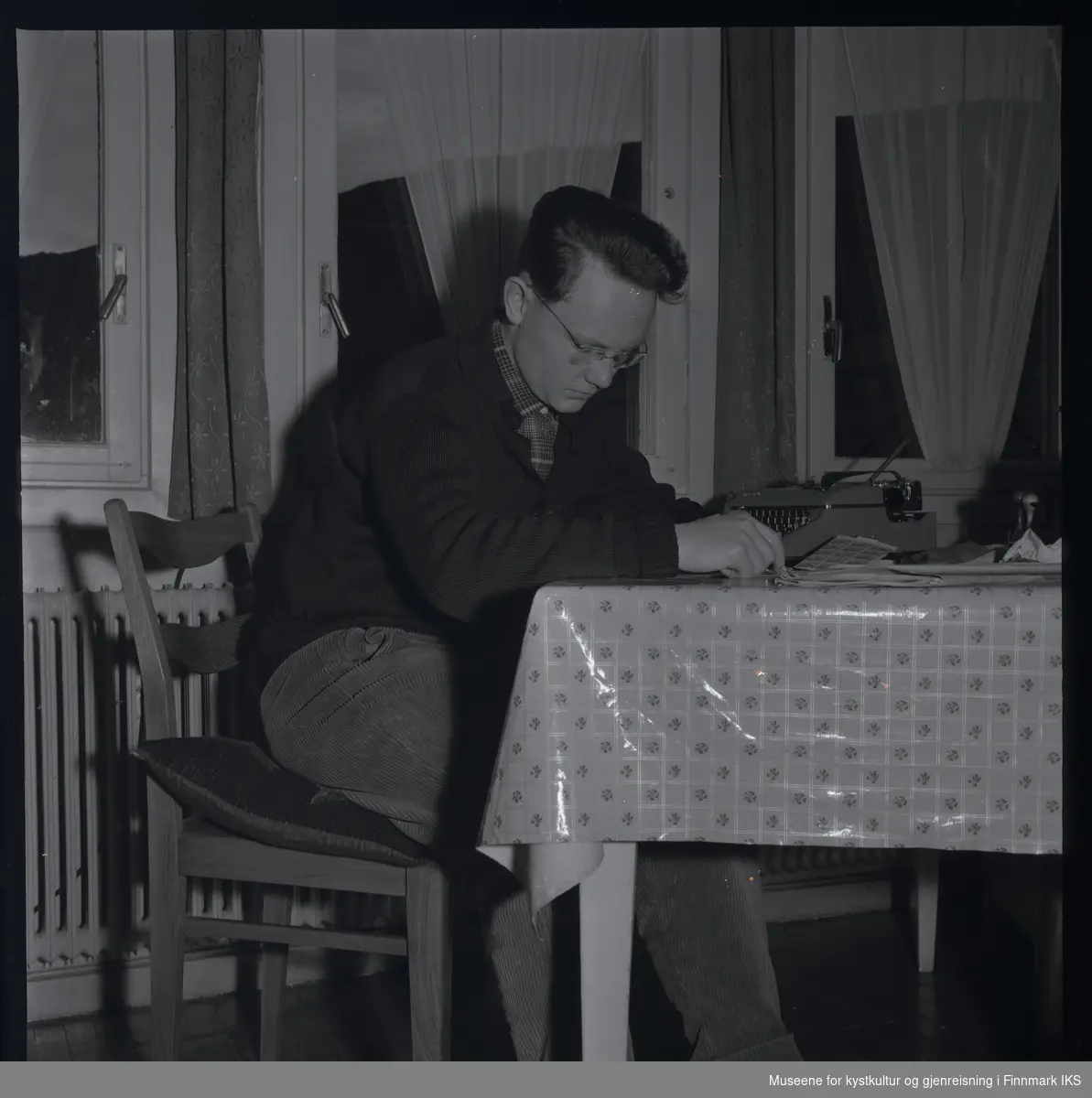 Friedrich Hörl sitter ved et bord og leser. På bordet, som er dekket med en voksduk, er det plassert en skrivemaskin, stempler og frimerker, samt en avis. Den unge mannen har en brille og er ikledd fløyelsbukse, rutet skjorte og strikket jakke.