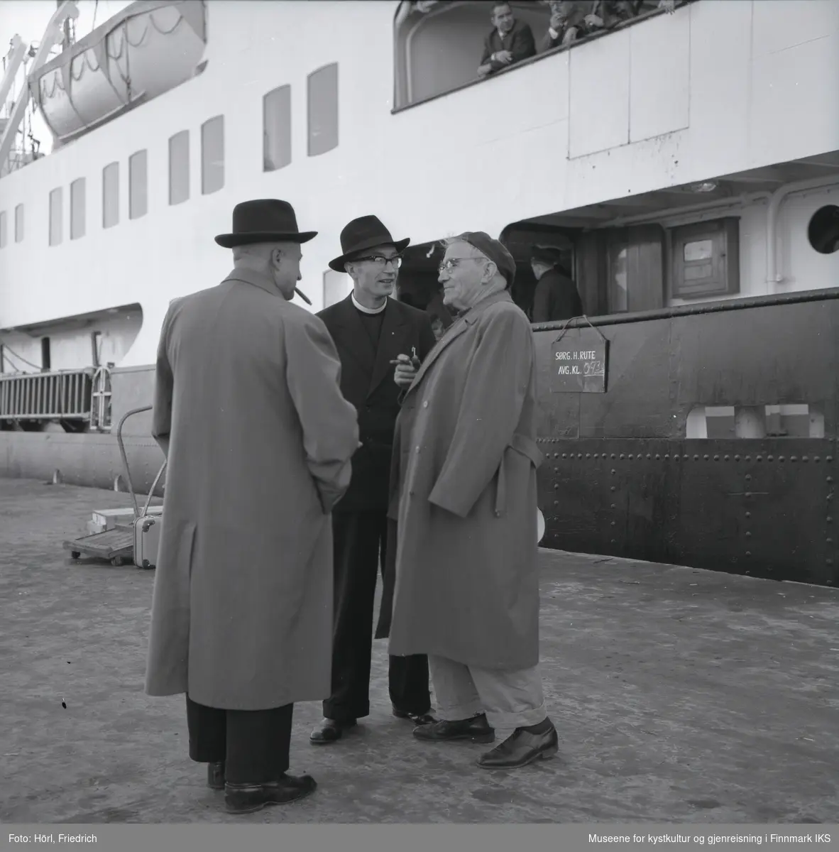 Tre men i samtale på kai i Harstad. To av dem tar røyk. I midten står Pater Spitz. En av de andre menn er Pater Rusche. I bakgrunnen ligger en skip til kais. Kofferter og transportgods står ved landgangen og øverst i bildet ser man folk om bord på skipet og en redningsbåt.