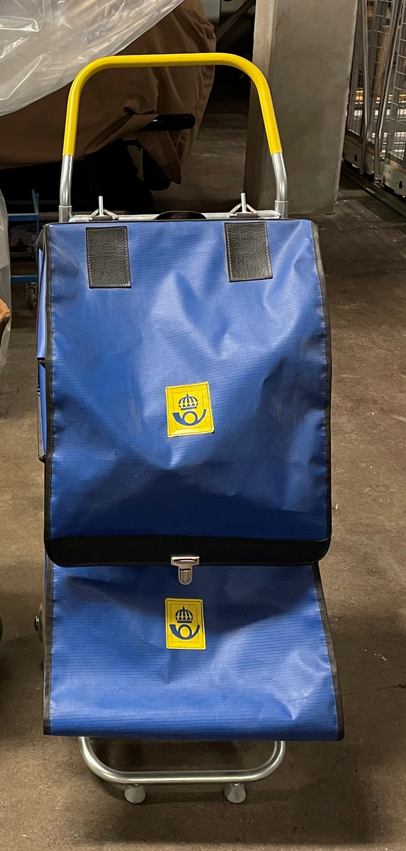 Brevbärarkärra i metall med två löstagbara väskor i mörkblå plastväv. Kärran har gult handtag och två hjul i gummi. Gul postlogotyp från perioden 1984-1990.