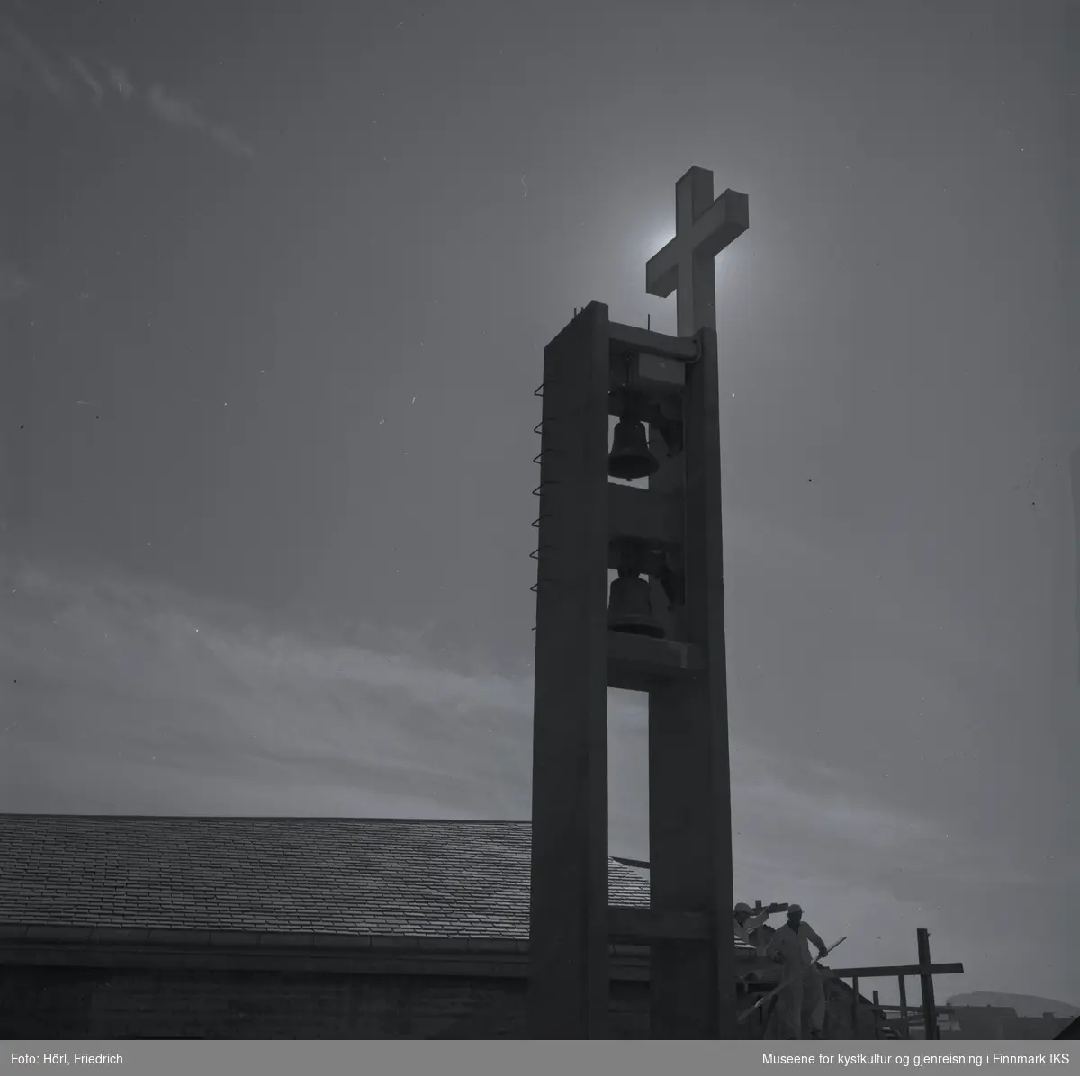 Bildet viser klokketårnet til den katolske kirka St. Mikael i Hammerfest i motlys av sola i 1958. Bildet er tatt fra sidebygget ut mot Sorøysundet der man ser Håja fjellet. Taket til kirka er ferdig bygd og belagt med skifer. På nedsida av kirka står det fortsatt et stillas og man kan se to arbeider der. I tårnet henger igjen de klokkene som allerede tilhørte til kirka før ødeleggelsene under andre verdenskrig. Det lyssatte korset er montert, men det er foreløpig kun sola som skinner.