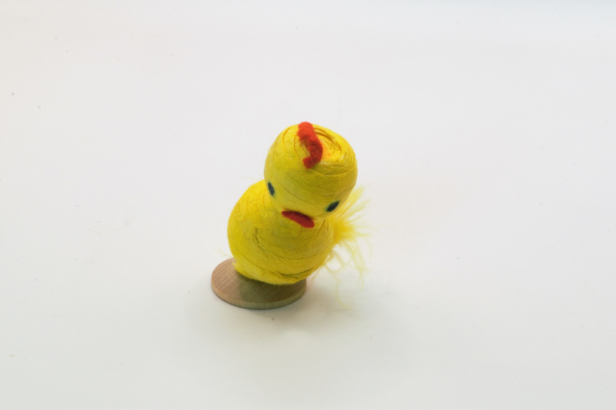 Kyllingfigur laget av gulmalte vattkuler, med nebb og hanekam av pålimt filt. Limt på sirkelformet støtte av tre.