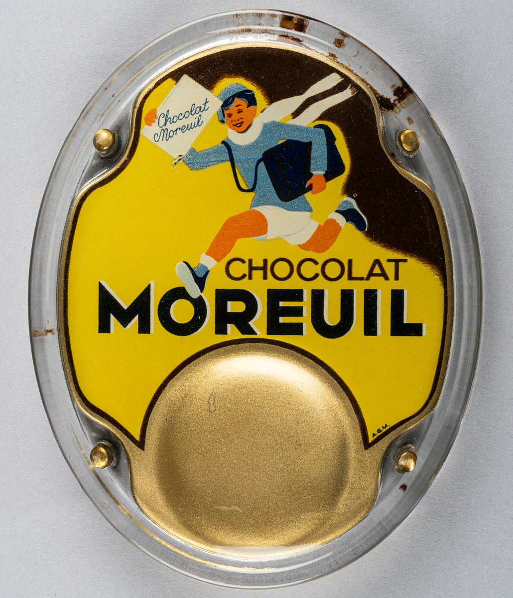 Myntbricka, oval, i glas, med guld, gul och svart bakgrund och springande pojke samt texten "CHOCOLAT MOREUIL" i svart.