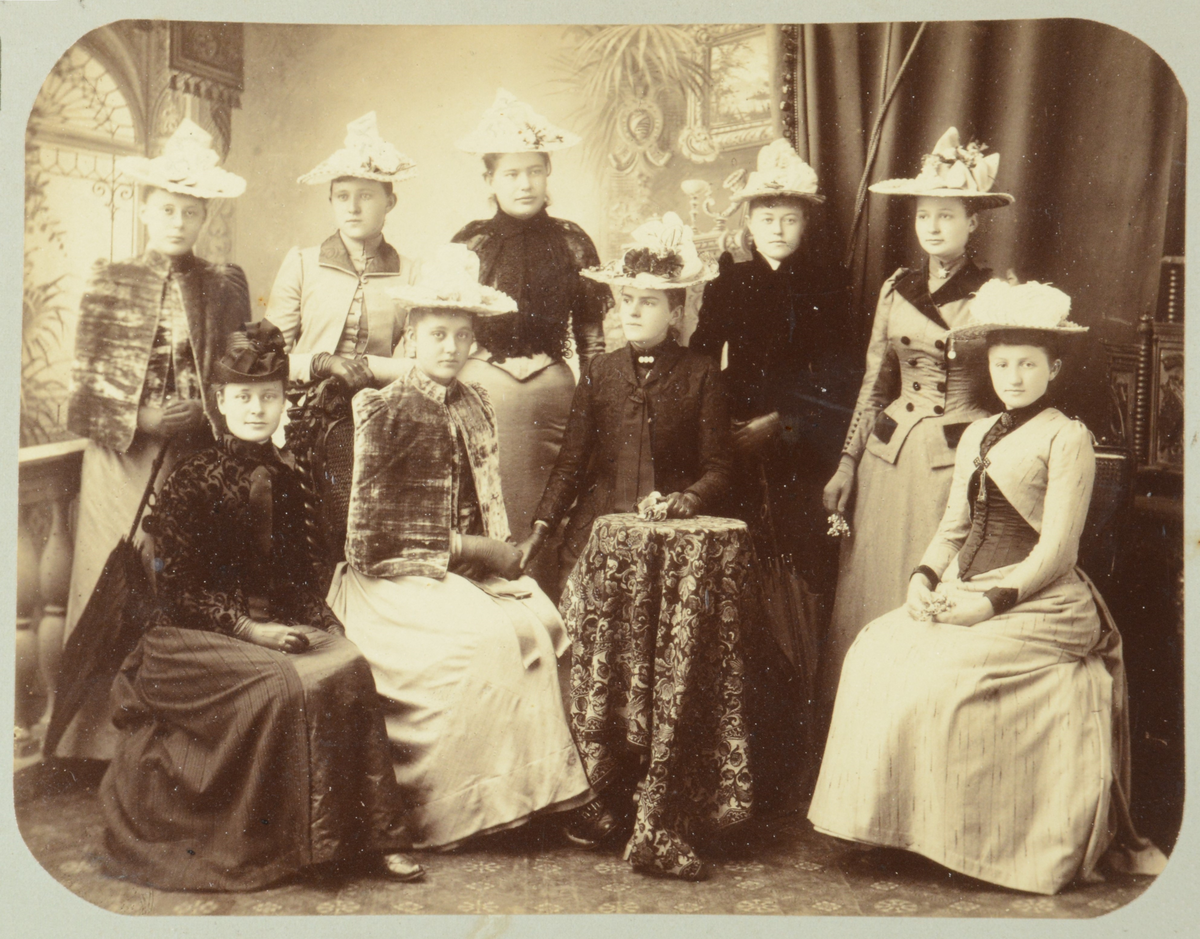 Ni unge kvinner med drakter og hatter stående og sittende rundt et bord med duk.
