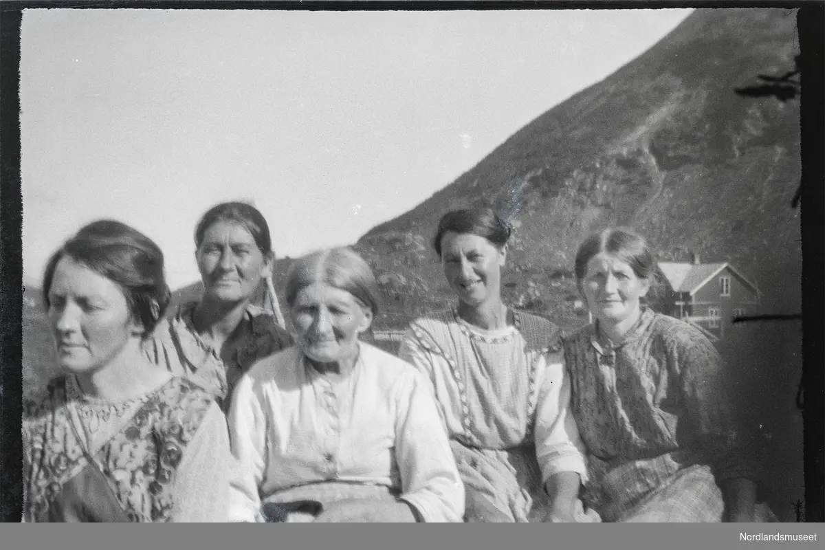 Gruppebilde av fem kvinner. Fra venstre: Anna f.1882 (usikker) , Arentine Karlsen, Marta Marie Andersdatter (f.1854, d.1929), Aminde og Regine /eller Anna Andorsdatter.  I bakgrunnene ses en bergrygg og et bolighus.