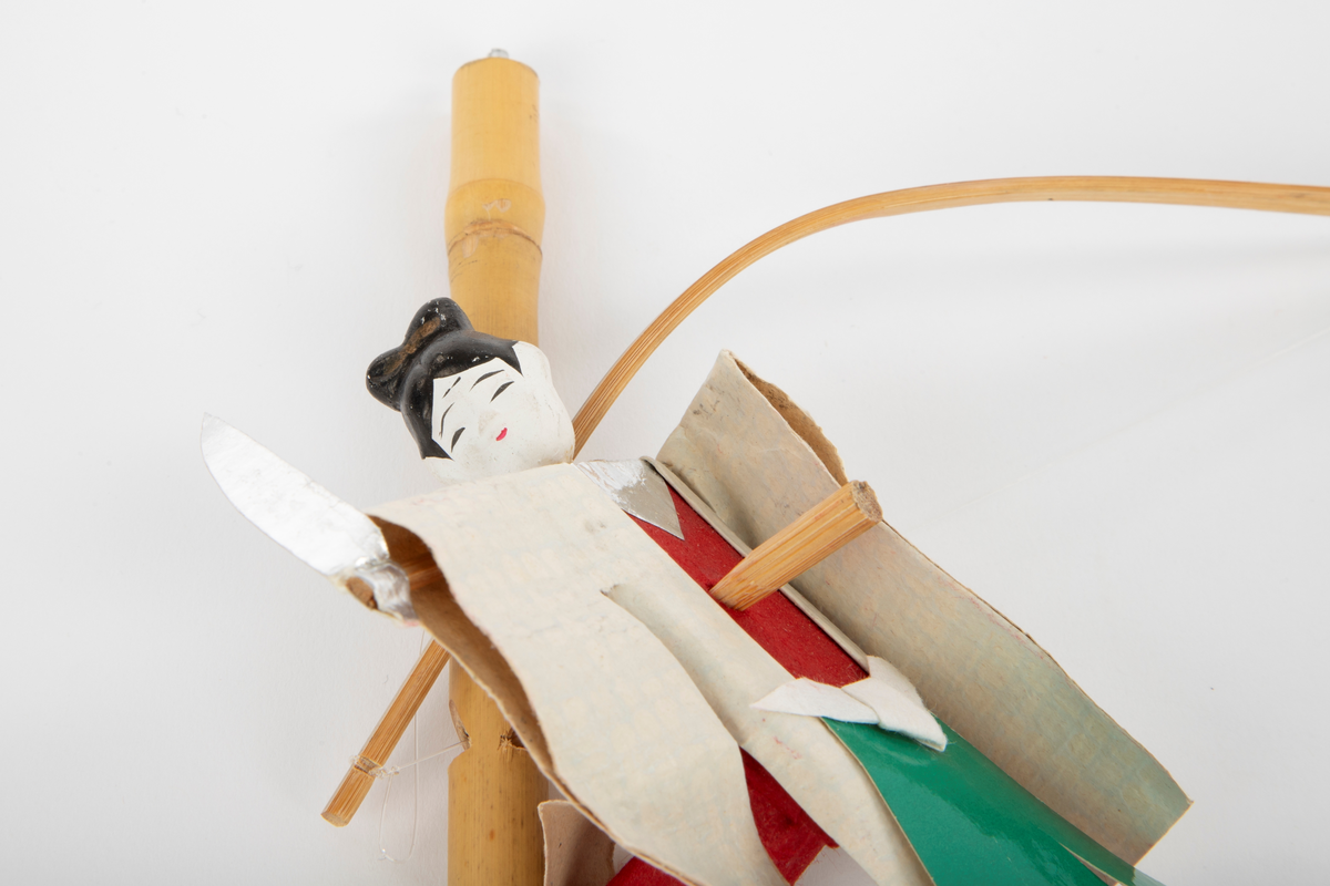 Bevegelig dukke i papir med trepinner til kropp og hode av gips, som forestiller den trofaste tjeneren til samuraien Yoshitsune. Dukken er hvitmalt med svart hår og har rød og hvit drakt med et grønt klede foran. Dukken holder i en sigd og har en bue, som kan henges på kroppen.