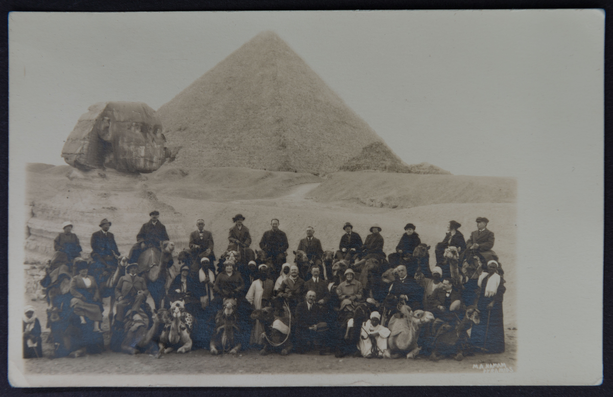 Gruppbild av turister i två rader som poserar framför en pyramid. Turisterna sitter på kameler. Den främre radens kameler hukar, den bakre stående. Möjligen är ledsagare stående bredvid.

Text kortets baksida "Vid Cheops pyramider och sfinxen nära Cairo 8/2 1924."