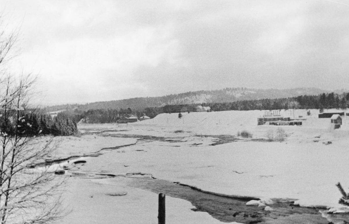 Området nedenfor terskeldammen som stuvet opp vann til kraftverket ved Skjefstadfossen i Heradsbygda i Elverum i Hedmark, fotografert fra den østre elvebredden tidlig i mars 1937.  Fotografiet er tatt fra elvebrinken på østsida av vassdraget, med kameraet vendt sørvestover, der den delen av elveløpet som gikk mot vestsida av Tjuvholmen var ei åpen, stilleflytende råk mens det øvrige elvefaret var is- og snødekt. Til høyre i bildet ser vi den vestre delen av kraftverksdammen med luker som kunne brukes fil å regulere vanntilførselen til rørene som førte vannet drøyt 400 meter langs vestre elvebredd ned til kraftstasjonen.  Fotografiet ble tatt på ei årstid da vannføringa i Glomma var svært beskjeden, og da de foregikk reparsjonsarbeider på sjølve kraftverksdammen, jfr. SJF.1992-05763.
