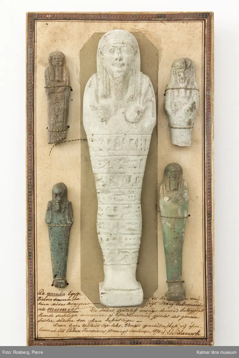 KLM 44649:2. Skulptur, gravfigur, 5 st. Den största i gråvit sten. Står på liten upphöjning. Hieroglyfer inristade från midjan och ner till fötterna. En av de mindre har även den hieroglyfer inristade. Två av de mindre är grönaktiga i färgen. De fyra mindre är fastmonterade med snöre på kartongskiva. Den större har lossnat. Text på kartongskivan: De gamla Egyptierna bevarade sina dödas kroppar så länge som möjligt. Den af balsamering och mumier. De dödas själslif ansågs dermed betryggadt. Kunde verkliga mumier ej åstadkommas, gjordes det genom bilder, sådana som ofvan befintliga.