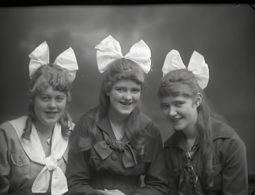 Ateljébild. Tre äldre flickor med stora rosetter i hår och ringning. Frisyrerna är omsorgsfullt lockade och arrangerade. Beställare: Anna Lill Reinholdsson, Falkenberg.