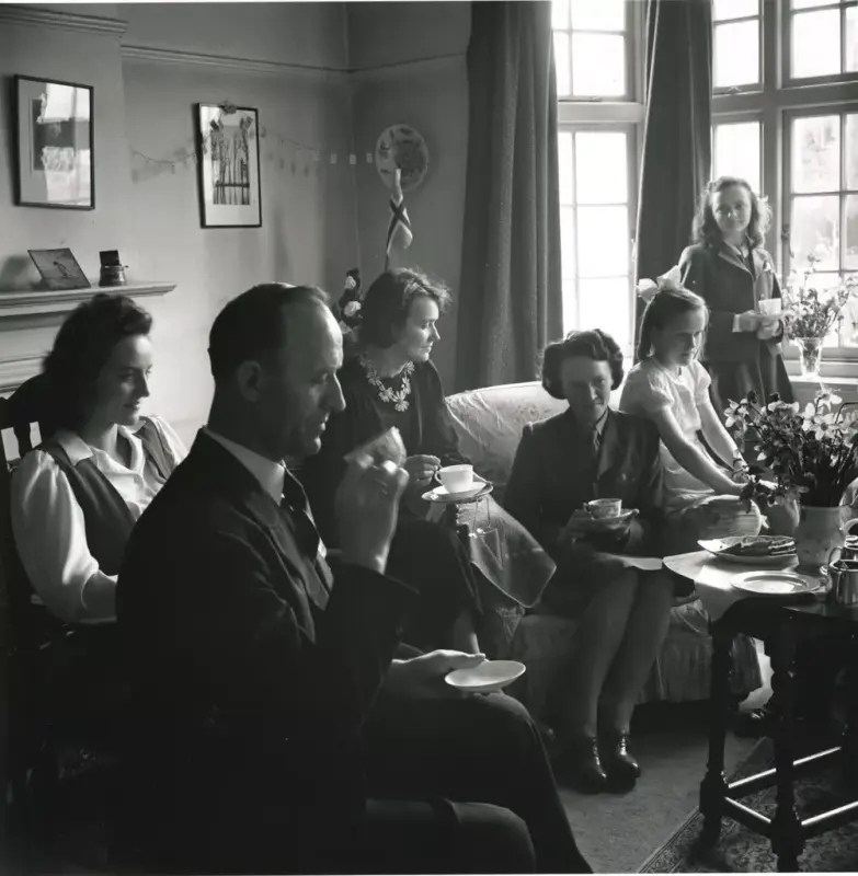 Ein familie drikker te i ei stue i London under krigen. Seks personar, ein mann tre kvinner og to jenter. Eit lite, norsk flagg i bakgrunnen.