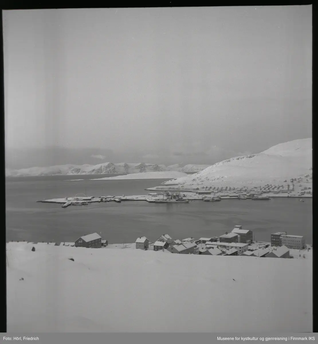 Bildet viser utsikten over Hammerfest og bydelen Fuglenes om vinteren. I forgrunnen ser man ulike bygg i sentrum og på Hauen, blant annet hotellet. I bildemidten ser man bydelen Fuglenes med slippen og et skip som ligger til kais. Takene er snødekket på gjenreisningshusene på Rosmolla og bak der, på Meland, står det fiskehjeller. I Sørøysundet ligger Melkøya og Sørøya lengst bak i bilde.