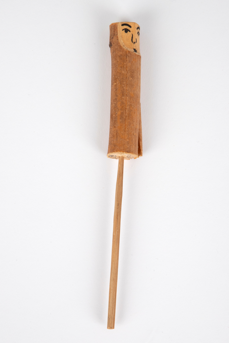 Tredukke laget av en trepinne med bark montert på en tynn pinne. Ansikt av ubehandlet tre, malt med svart. Dukken forestiller en kvinne og hører sammen med NK1991-029-004 (mann).