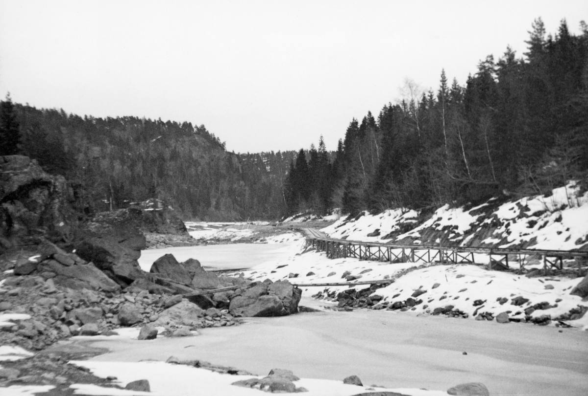 Fra bygginga av en ny dam ved Sølvstufossen, øverst i Ågårdselva i Tune i Østfold, som er en del av Nedre Glommas vestre løp. Anleggsarbeidet startet vinteren 1936, i en periode da det knapt rant vann i elveløpet. Fotografiet viser elveløpet i medstrøms retning. Langs den nordre elvebredden (til venstre på bildet) hadde man bygd en trallebane på trebukker, antakelig for å kunne transportere materialer og utstyr noenlunde effektivt til og fra anleggsområdet. Ågårdselva er et cirka fem og en halv kilometer langt vassdrag som renner fra den nordvestre enden av Isnesfjorden (Vestvannet) i Nedre Glommas vestre løp, sørvestover gjennem berglendt terreng mot innsjøen Visterflo. Høydeforskjellen mellom Isnesfjorden og Visterflo er på bortimot 25 meter. Ågårdselva har tre fossefall, det øverste her ved Sølvstu, deretter ved Valbrekke og nederst ved Solli. Behovet for den dambygginga vi ser på dette fotografiet var forårsaket av selskapene Borregaards og Hafslunds inngrep i Glommas østre løp, hovedløpet, som i lavvannsperioder gjorde det svært vanskelig å få tømmer som skulle til bedrifter nedenfor Sarpsfossen via Mingevannet, Isnesfjorden og tømmertunnelen til Eidet gjennom det trange sundet ved Trøsken. Dette forsøkte man å løse ved å heve vannspeilet i Isnesfjorden med en dam som skulle plasseres 10-15 meter ovenfor den eksisterende dammen ved Sølvstufossen. Se mer informasjon under fanen «Opplysninger».