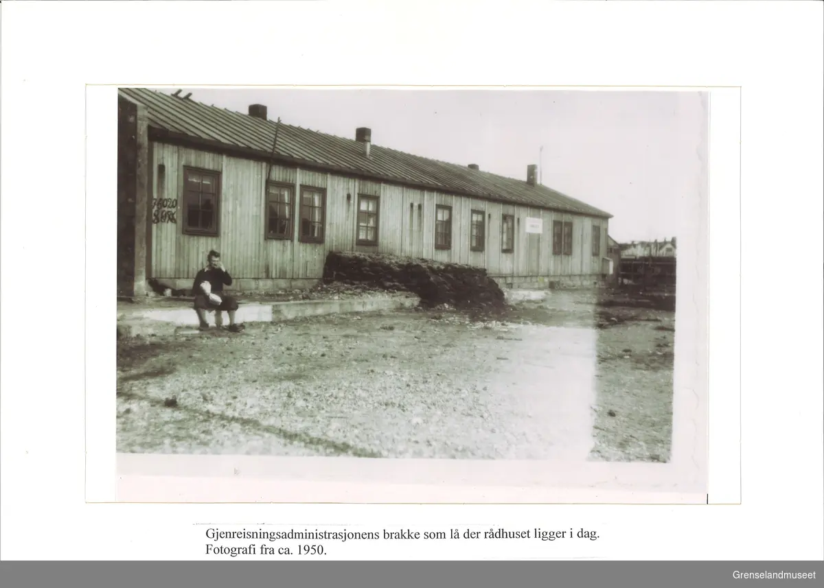 Avbildet i svart-hvitt er gjenreisningsadministrasjonens brakke i Kirkenes som lå der rådhuset ligger i dag. En person sitter på fortauet foran brakkebygget.