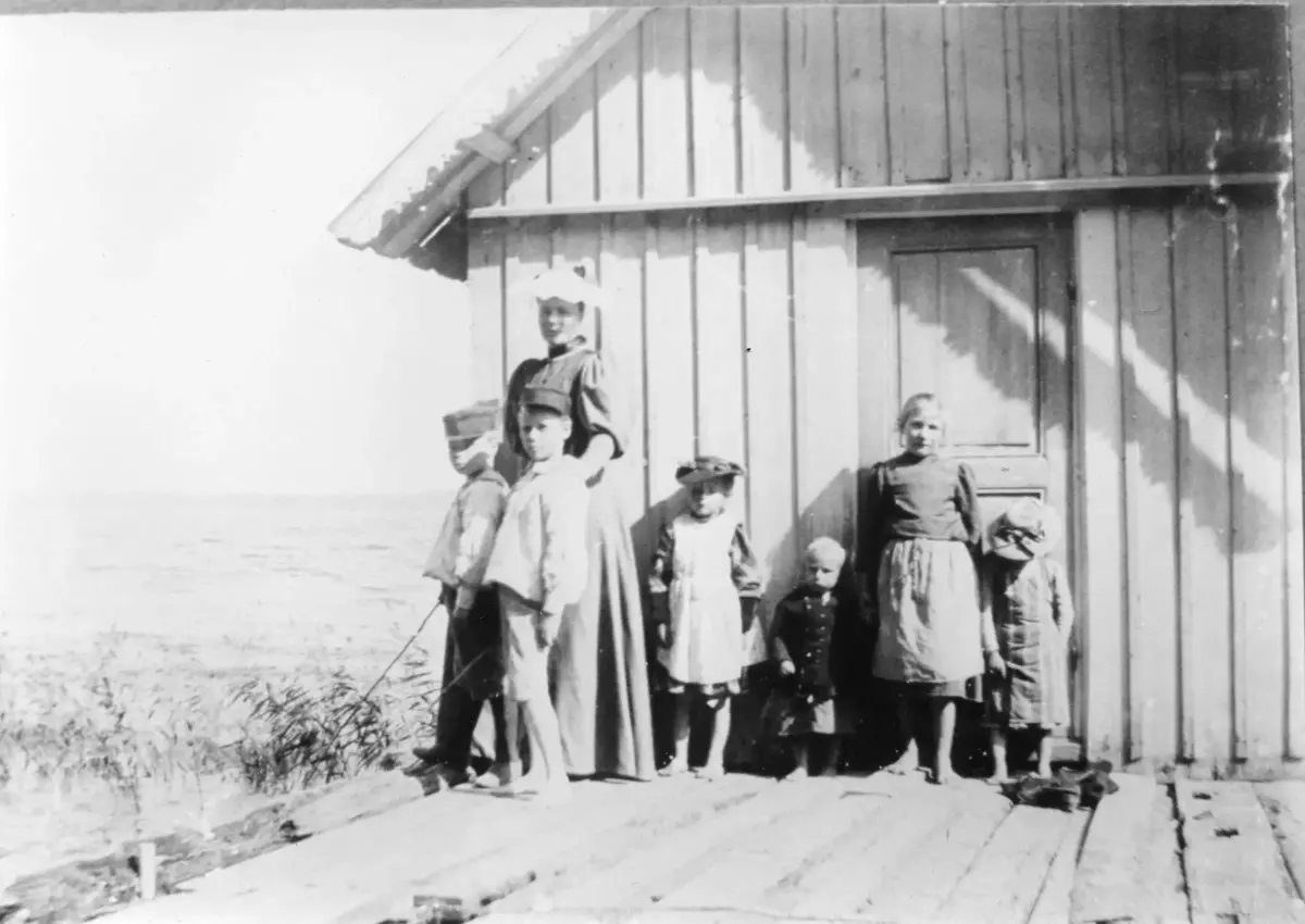Damen till vänster är Nanna Salmonson, närmast framför henne står sönerna Carl och Otto. Övriga personer okända. Solar sig vid sjöbodsväggen. I bakgrunden Våmfjärden.