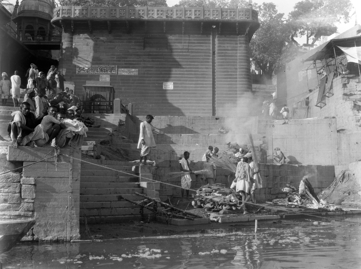 Likbål ved bredden av Ganges. Menn i trappen ned til elven. I bakgrunnen en bygning, Benares, India.