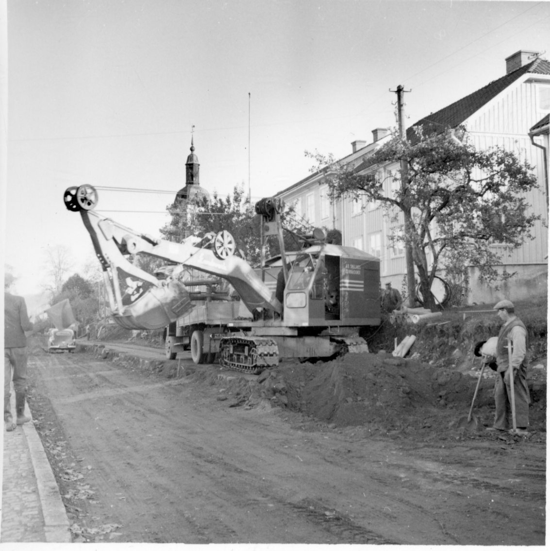 Fyra män, varav en flaggvakt, står vid en grävmaskin i arbete. En lastbil står bortom grävmaskinen. Södra delen av Brahegatan i Gränna breddas.