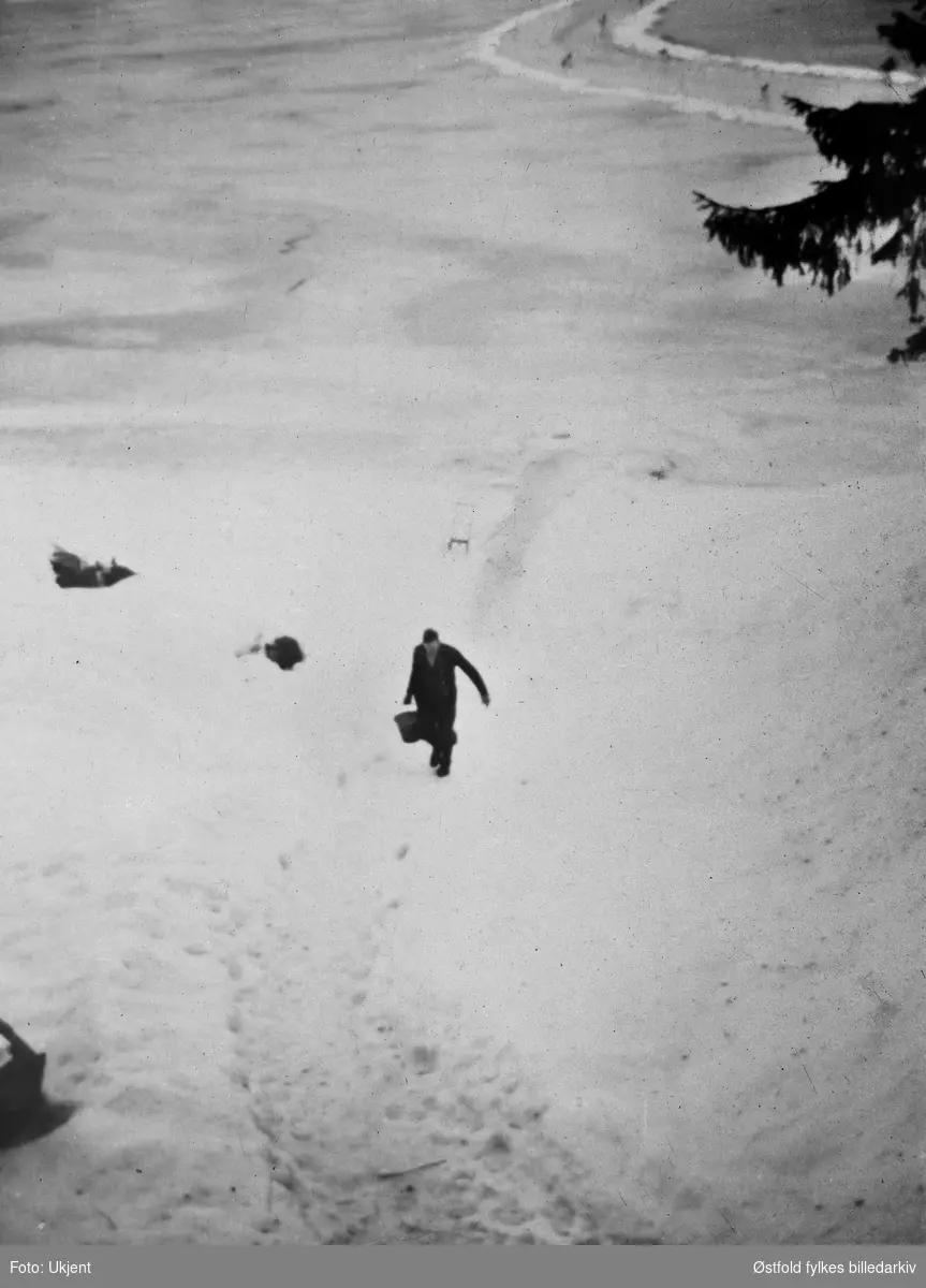Vinterveien over Glomma, Grønnsund i Skiptvet, ca. 1925. En sparkstøtting og en person som bærer en bøtte i forgrunnen.
På fotokort er personen på bildet oppgitt til å være Marie Børud, Høgåstangen. Usikker opplysning, personen på bildet ser ut til å være en mann.