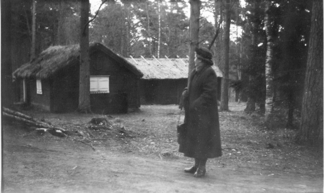 En kvinna med hatt, kappa och handväska tittar mot en gammal enkelstuga och ekonomibyggnad med halmtak.