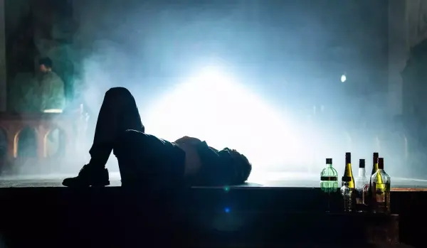 Silhuetten av en mann som ligger på ryggen med knærne opp og en samling flasker med alkohol bak hodet. Hvitt lys i bakgrunnen i et ellers mørkt rom.
