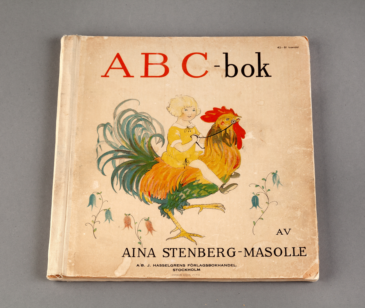 ABC-bok av Aina Stenberg-Masolle. Text och illustrationer för alla bokstäver i alfabetet. Hårda pärmar. På framsidan färrik illustration med ett barn sittandes på en tupp och hållandes i tömmar . Under och intill olika blommor.  På insidan av första uppslaget står hanskrivet. "Till Margaretha från mormor o morfar Julen 1947"