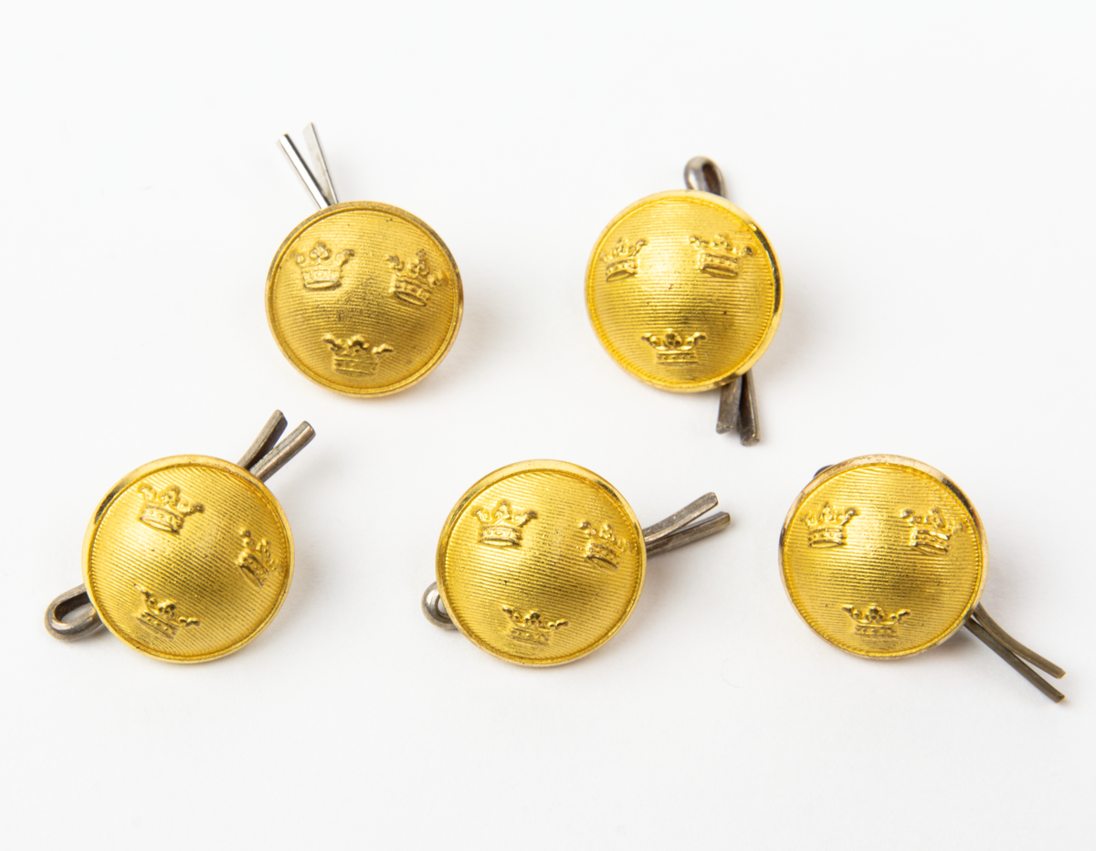 Guldfärgade räfflade knappar med tre kronor. På baksidan sitter en saxsprint instucken i öglan.
En av knapparna är tillverkade av Lagerströms Fabriks AB Mjölby och fyra av C.C. Sporrong AB Stockholm.