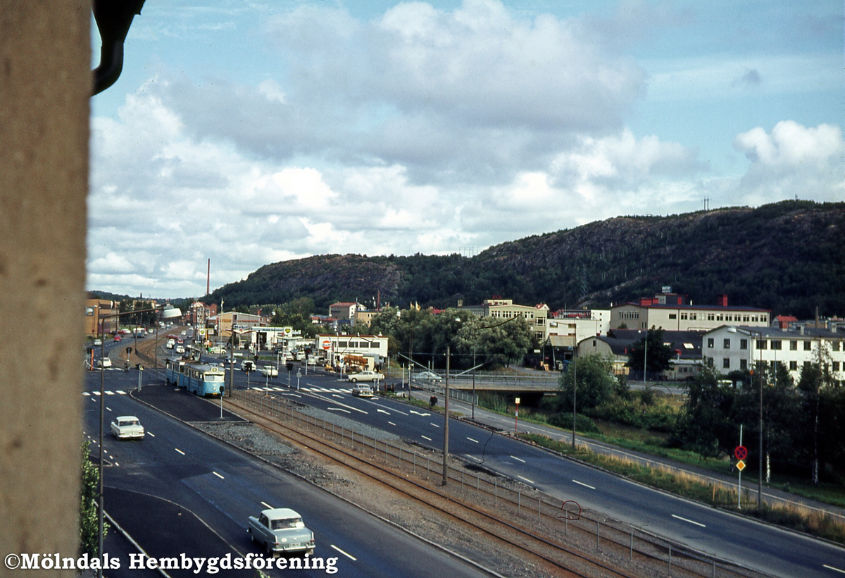 Trafik på Göteborgsvägen och Lackarebäcksbro i Mölndal, den 3/9 1967. Sista dagen med vänstertrafik. Lackarebäcksbron går över Mölndalsån. Till vänster ses 4:ans spårvagn komma. I bakgrunden ses bebyggelse i Lackarebäcks industriområde samt Lackarebäcksfjället och Rallarberget.