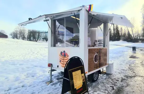 Liten serveringsvogn med oppslåtte salgsluker i to retninger og columbiansk flass på hjørnet står på snøen med en plakatbukk med gul prisplakat foran og Hamardomen i bakgrunnen.