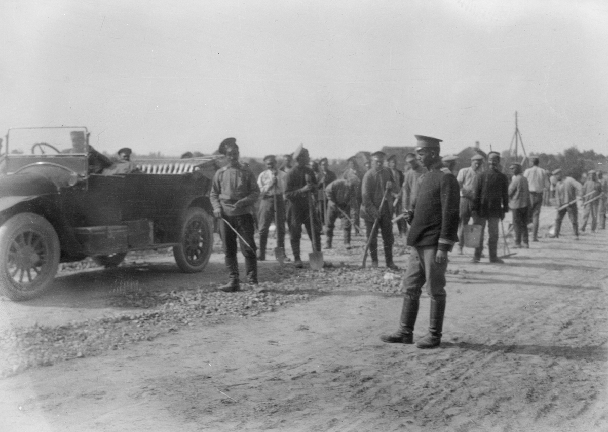 En stor samling män jobbar för hand med väganläggning i Centraleuropa under första världskriget. Utrustade med krattor och spadar. En bil står i förgrunden. Tillhörande Österrike-Ungern.