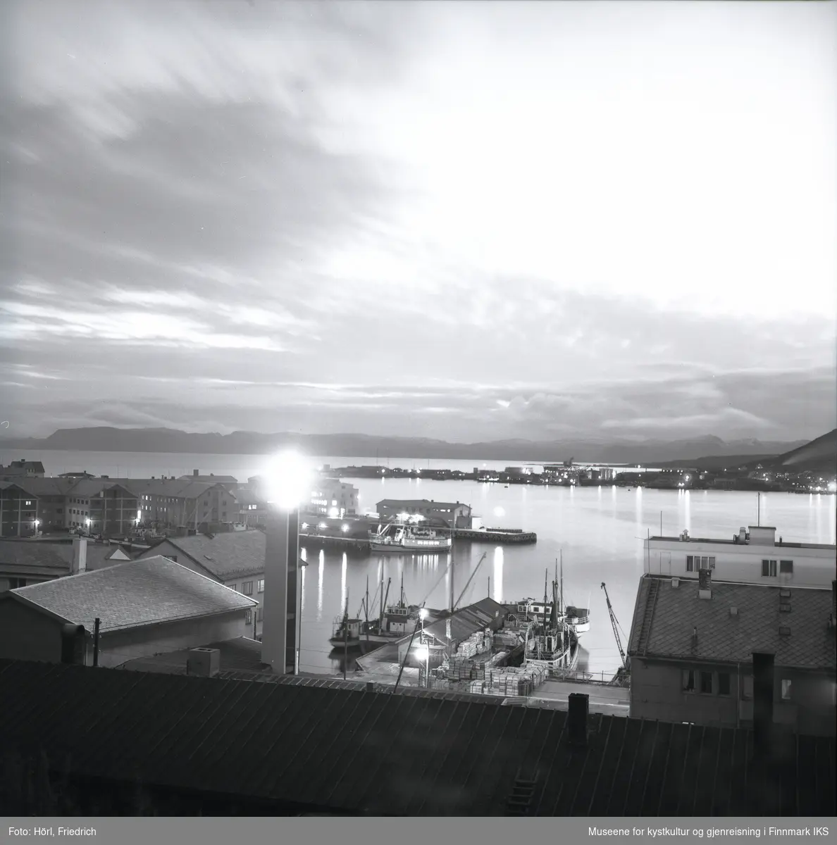 Det er skumringstime i Hammerfest og fotografen tar et bilde fra fjellsiden av Salen ut mot havna. I forgrunnen ser man et brakketak. En lys flekk på bildet er der det lyssatte korset ved klokketårnet til den nybygde katolske kirka St. Mikael er. Gatelysene er på. Ved kaia ligger det båter og på andre siden av bukta ser man bydelen Fuglenes. Lengst bak i bildet ser man silhuetten av Sørøya.