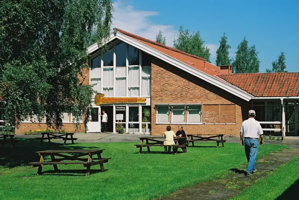 Murbygning med høye vinduer i gavlveggen. Døra nedenfor holdes åpen av en mann. Skilt over døren har inskripsjon Norsk utvandrermuseum og Norwegian Emigrant Museum. Gressplen i forgrunnen med benker og noen besøkende. Bjørketre til venstre.