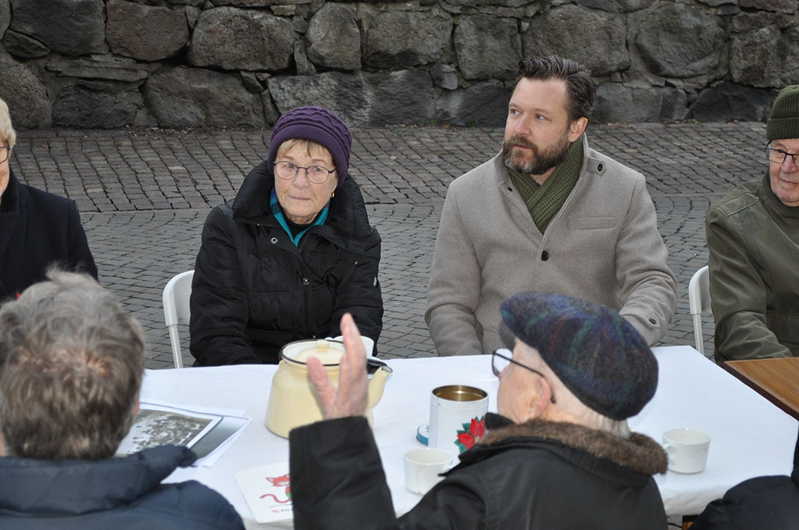 Män och kvinnor sitter vid ett dukat bord på Brahegatan i Gränna. Lyssnar på en man med keps som pratar.