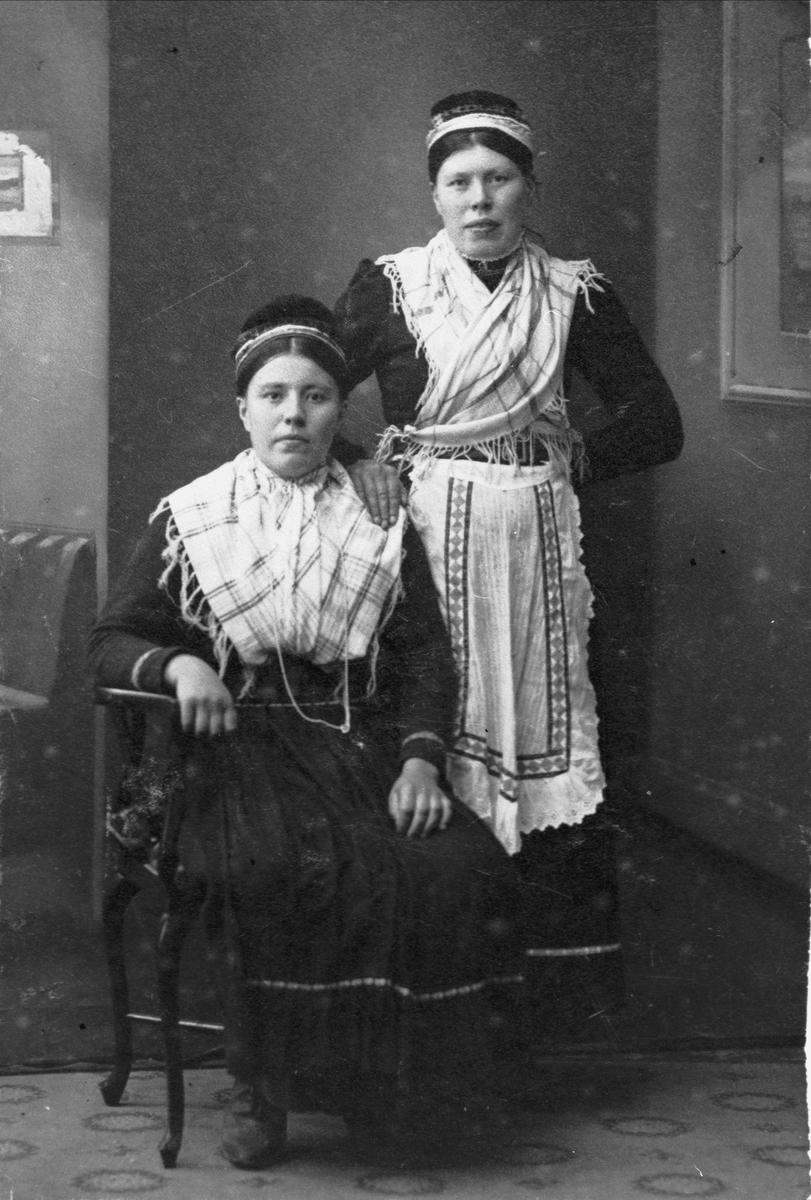 Studioportrett av to unge kvinner i samisk drakt. Den ene sitter i en stol, den andre står bak.