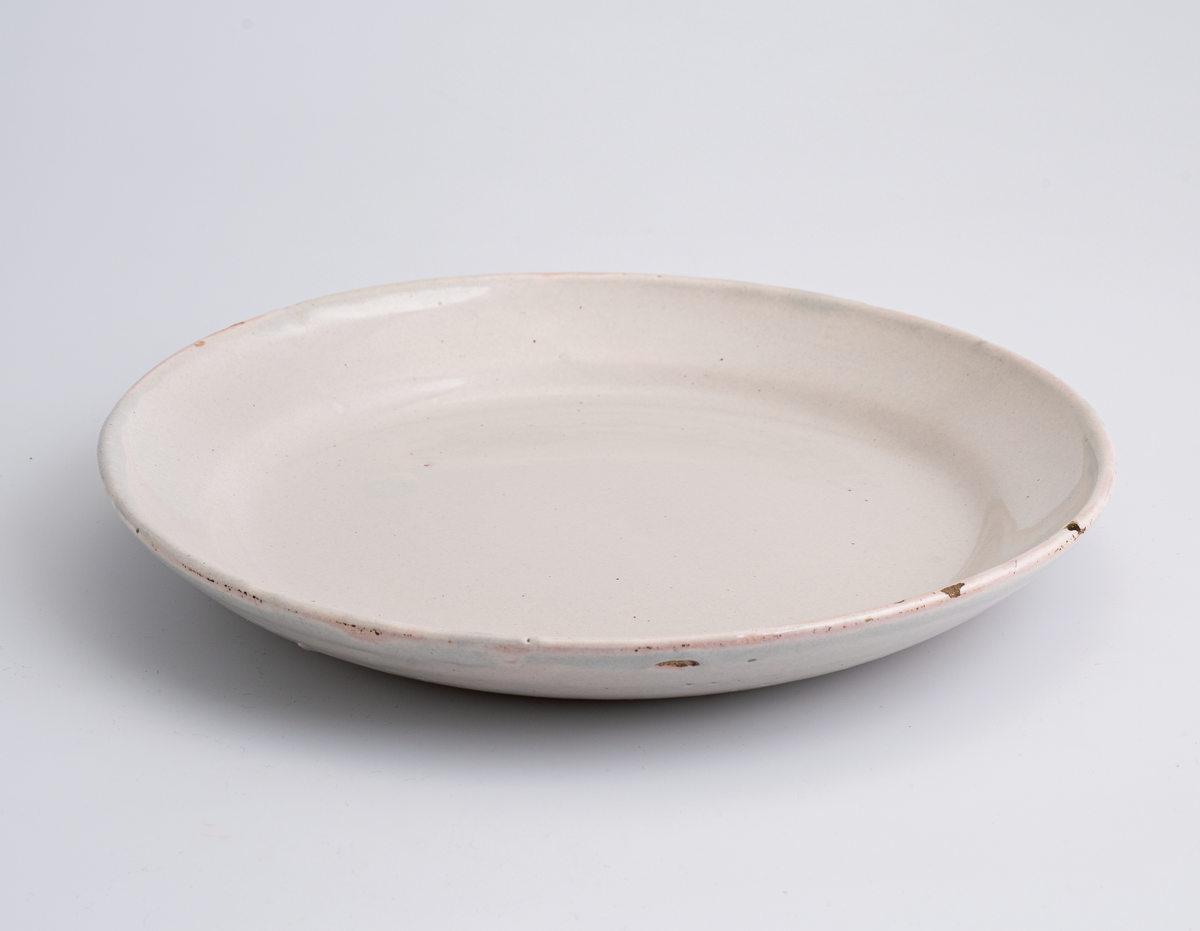 En flat tallerken av keramikk/porselen. Den har bly- og tinnglasur og er hvit på farge. Tallerkenen er rund med en relativt høy og skrånende kant. Den har ikke dekor utover glasuren. Tallerkenen er en av 11 identiske tallerkener som trolig er fra tidlig på 1700-tallet.