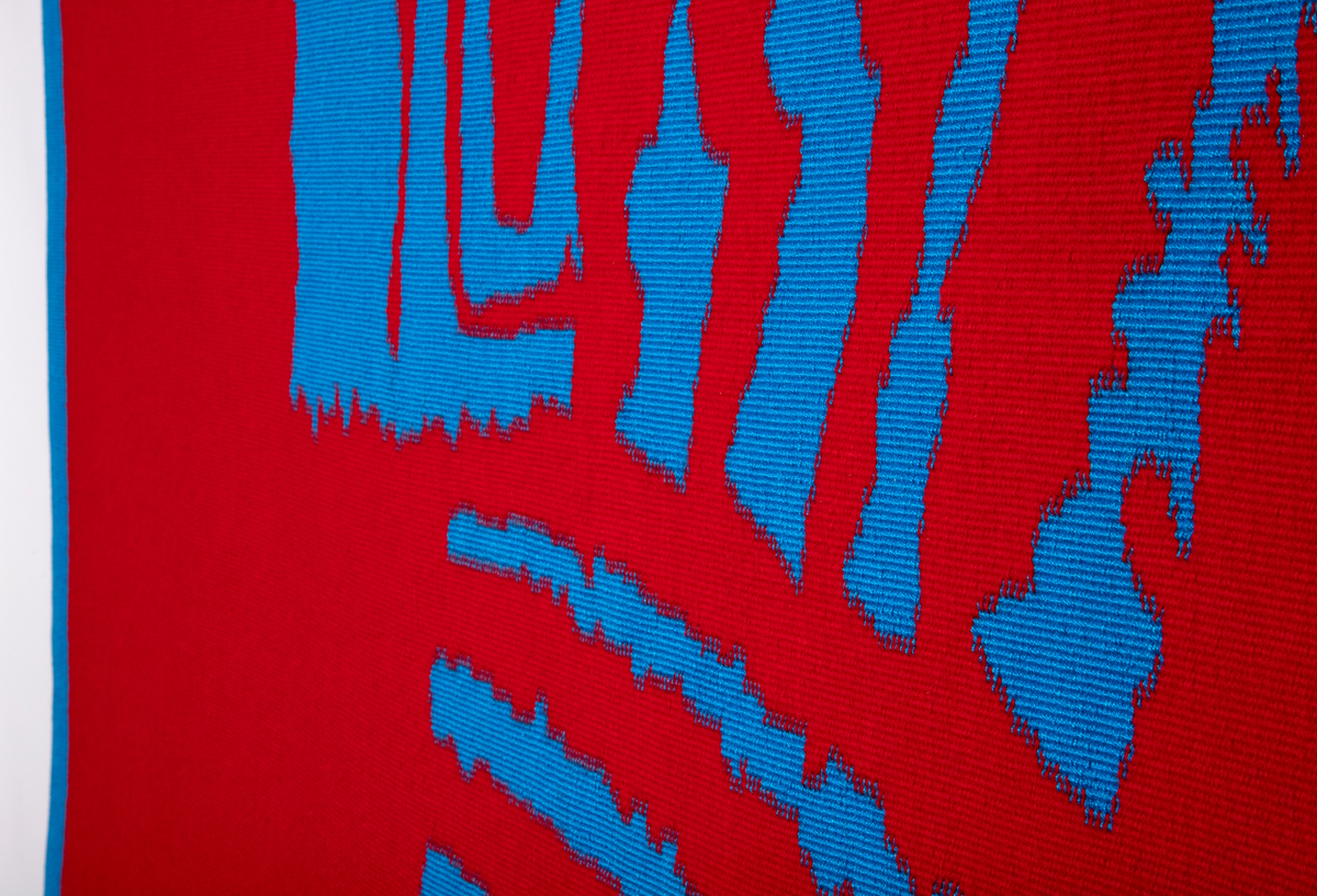 Rektangulær billedved i rødt og blått. Portrettet viser ansikt og hals bygget opp av rette og vinklede former i blå. Formene har ujevne kanter. Bakgrunnen er rød, inrammet av et blått felt. Figuren er plasert i øvre høyre hjørne. Bunnen er i ull, og mønster i silke.
